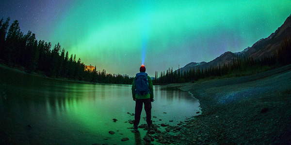 Homme debout sur la rive d’un lac, observant le ciel nocturne à la lueur de sa lampe frontale.