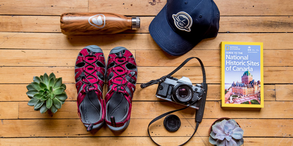 An arrangement of gear including a water bottle, cap, sandals, camera and travel guide.