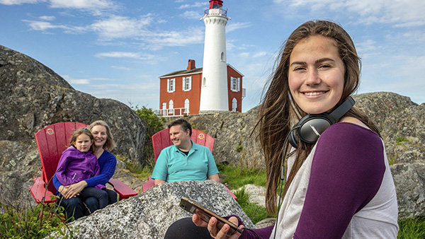 Une adolescente écoute de la musique pendant que sa famille est assise dans les chaises rouges de Parcs Canada au lieu historique national du Phare-de-Fisgard.