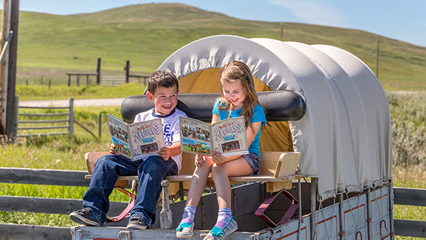 Two children sit on a chuckwagon as they check out the next challenge in their Xplorers activity booklet at Bar U Ranch National Historic Site.