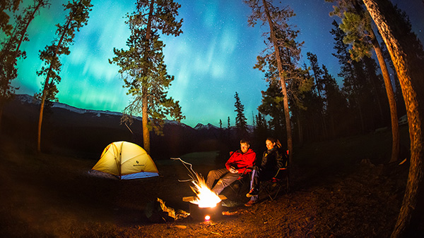 Two campers enjoying a campfire with northern lights in the sky.