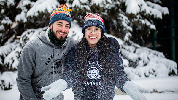 A young couple smiles at the camera and side-hug in their official merchandise in a snowy park.