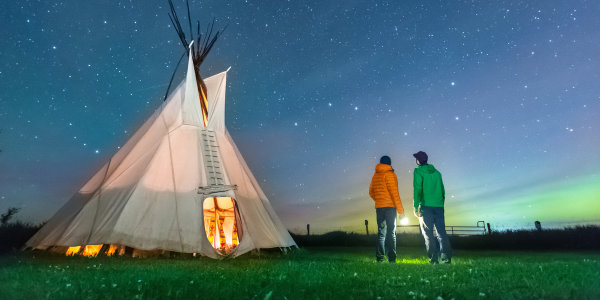 Two visitors gaze at the Big Dipper outside a tipi on a starry night at Rocky Mountain House National Historic Site.