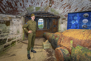 A tour guide dressed in WWII uniform stands beside historic artillery in the underground lower battery.